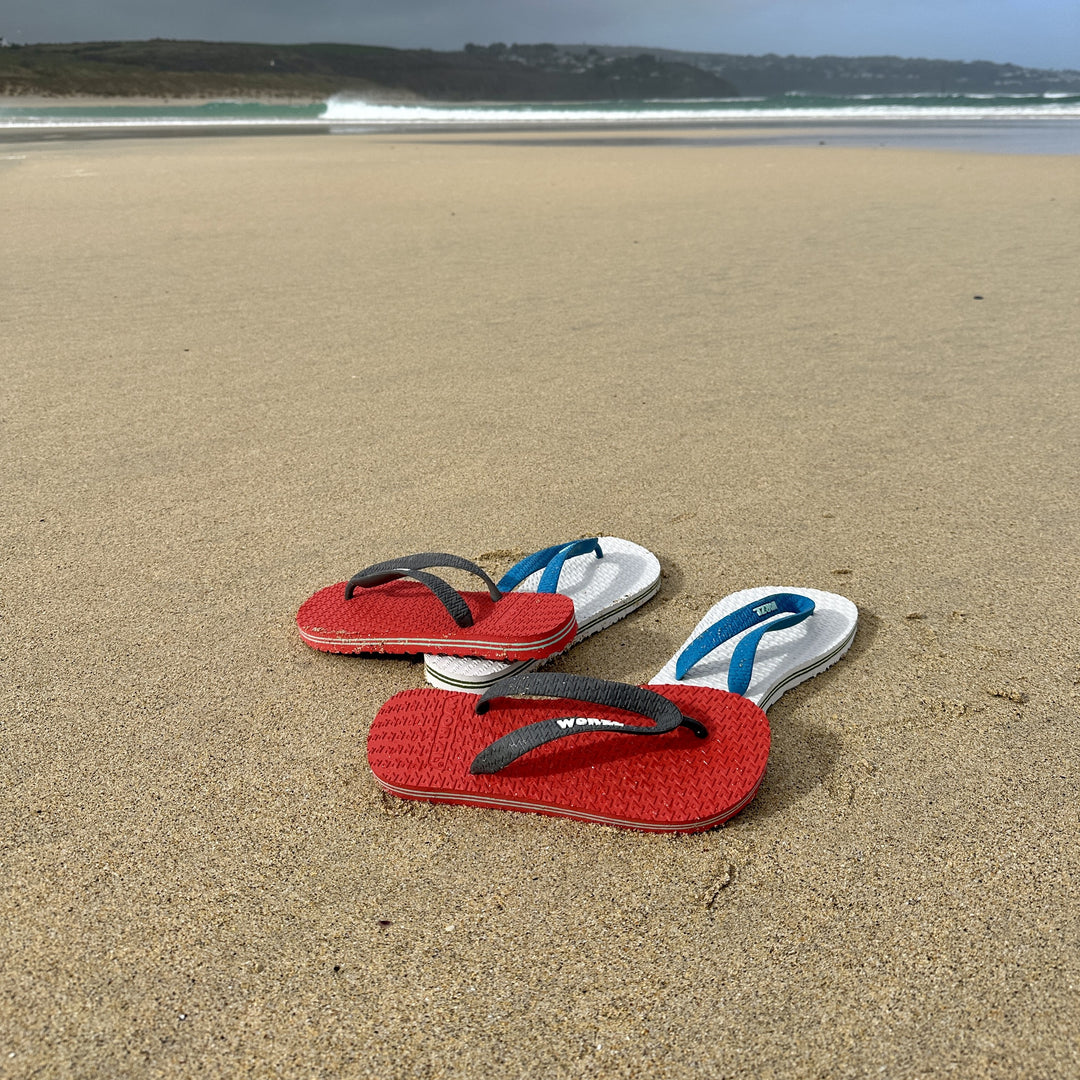 2 pairs of Worzl eco friendly flip flops on a beach by the sea.  Red with a grey strap and white with a blue strap.